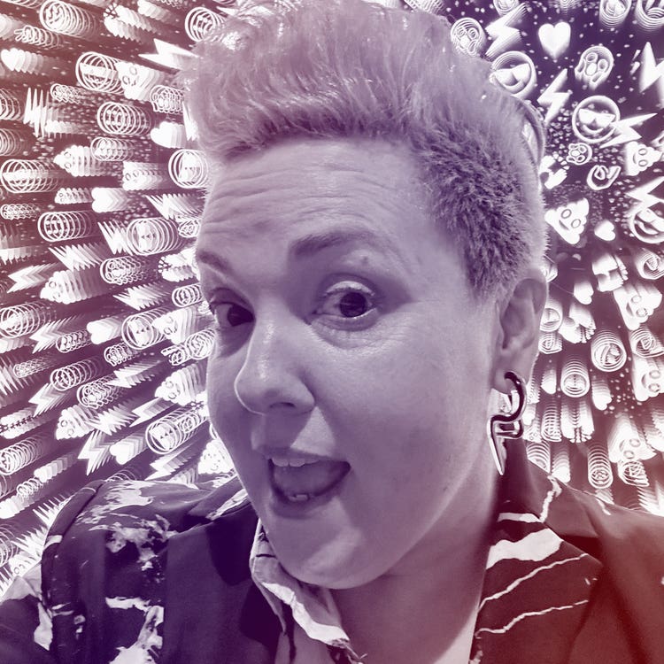 A photograph of a white woman with short blonde hair against a backdrop of digital illustrations. She's wearing a black-and-white blazer, black earrings and is interacting playfully with the camera.