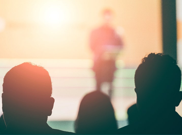 An out-of-focus photograh of the shadowed forms of the backs of three heads facing the blurred image of someone speaking from a stage.