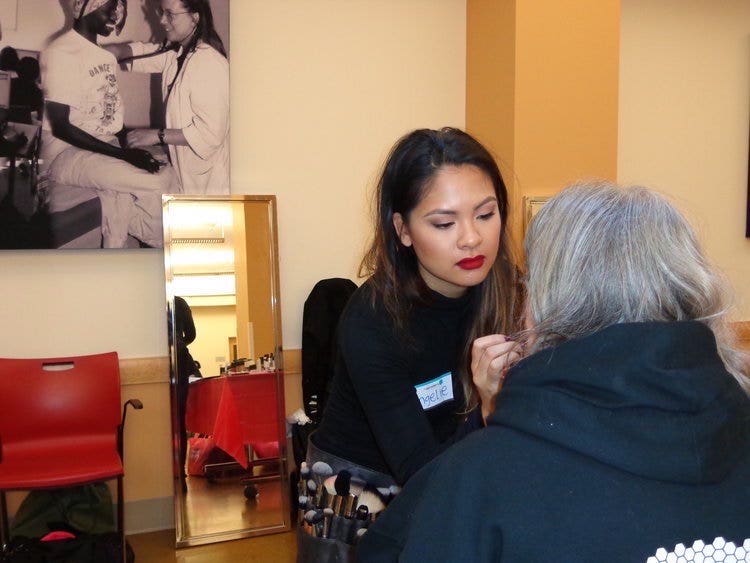 A photograph of two women. The woman facing the camera is applying makeup to the woman whose back is to the camera.