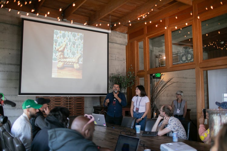A photograph of a white man (wearing a dark shirt and pants and holding a microphone) and a white woman (wearing jeans, and a white T-shirt). Both of them are facing an audience (whose heads are visible in the foreground) the man is speaking and behind them is large presentation screen with a digital image of dog-shaped couch against a patterned wall.
