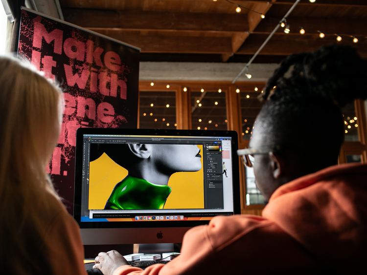 A photograph of two people (a woman with blonde hair and. a black man with glasses) with their backs to the camera, are looking at a computer screen. On the screen is a partial profile of a woman's neck and face against a mustard yellow background. Her face and hair (in a high bun) are in greyscale and her clothing (a green high-necked tunic) is in full color.