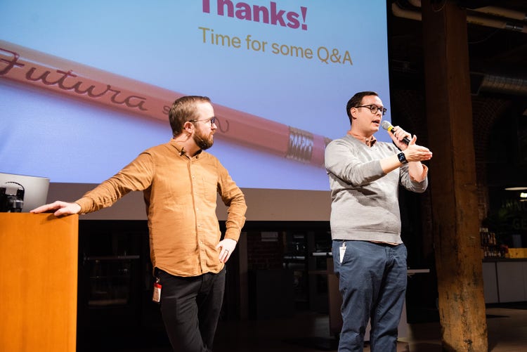 Two white men standing side-by-side on a stage. The man on the left is leaning on a podium, the man on the right is speaking into a microphone.