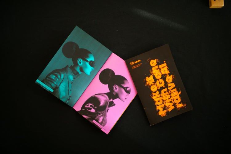 A photograph of two interior pages (left) and the cover (right) of a zine. The interior pages are of the same image, printed in grayscale on green (left) and pink (right) paper. The image is a woman in profile, wearing dark glasses, a high-necked tunic, and her hair in a high bun. The cover, also printed in greyscale (on orange paper) reads Make It with Generative AI.