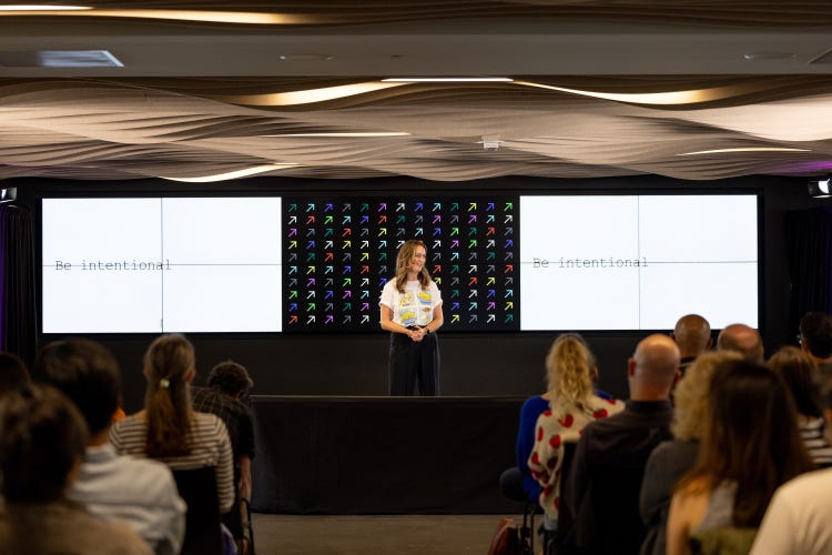 A white woman with shoulder length dark hair stands on stage in front of an audience, with a large screen divided into three sections behind her. The left and right sections display the phrase "Be intentional" while the center shows a grid of colorful arrows pointing diagonally upward to the right.