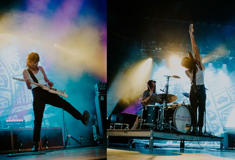 Two photographs of the same white woman with her dark hair styled in a modified mullet. On the left, she's wearing a white tank top, dark pants, and boots and is playing a bass guitar strapped over her shoulder. She has one foot in the air and behind her is a row of amplifiers, a graffitied wall, and stage lights casting yellow, purple, pink, and blue light onto a smoky background. On the right, the same woman is standing in front of a white man with long dark hair wearing a baseball cap and a T-shirt seated at a drum set with the words Tell Me How You Really Feel stamped on it. He's holding a drum stick in his right hand and looking up at her as she holds her bass guitar straight up. They're both on a very small stage against a background of stage lights casting an amber and blue hue on a graffitied wall.