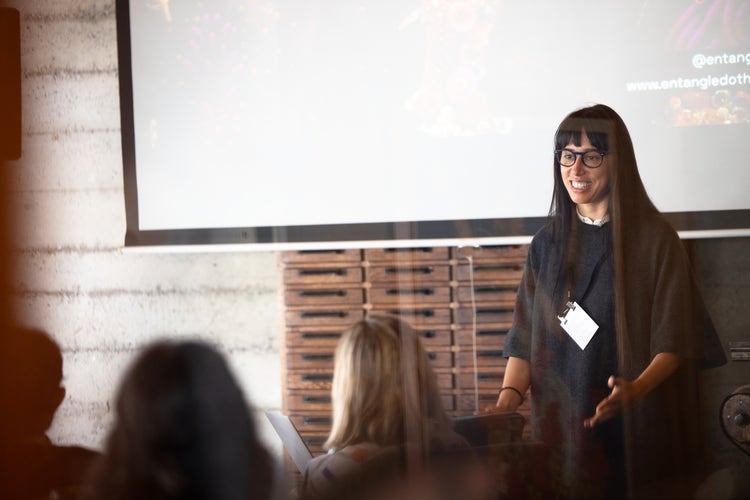 A photograph of a woman with long dark hair and bangs wearing glasses and a black top. She's smiling and standing with her back to a presentation screen. In the foreground are the backs of the heads of the people in the audience.