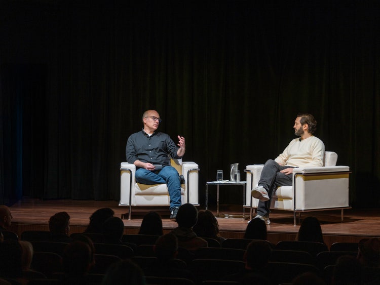 Two men in conversation are seated on a stage, in modern armchairs, against a backdrop of black curtains. The man on the left is wearing a dark shirt and blue jeans and the man on the right a light-colored sweater and black jeans. Between them is a small table holding a pitcher and two glasses of water. The backs of the heads of the audience are visible in the foreground.