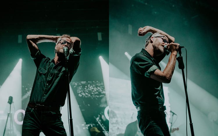 Two photographs of the same white man with short blonde hair wearing glasses, a plaid button-down shirt, and jeans. In both images he's singing into a microphone on a stand but in the image on the left he has both hands behind his head and in the image on the right he has his left hand behind his head and his right hand on the microphone. The stage lights are creating a bright white light against a green background.