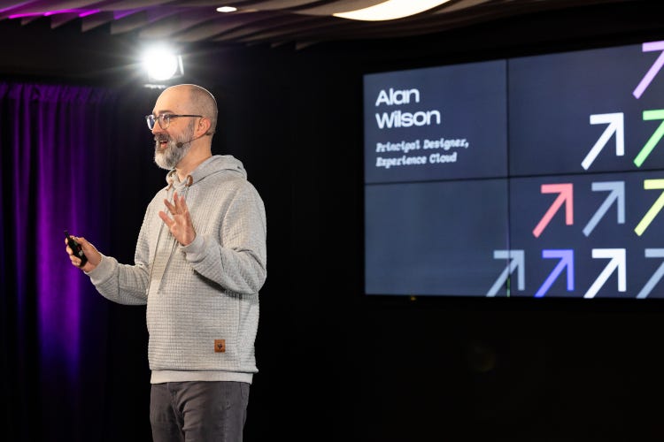 A white man with glasses stands on stage holding a device in one hand and gesturing with the other. Behind him, a large screen displays his name and job title (Alan Wilson, Principal Designer for Experience Cloud) alongside a grid of colorful arrows pointing diagonally upward to the right.