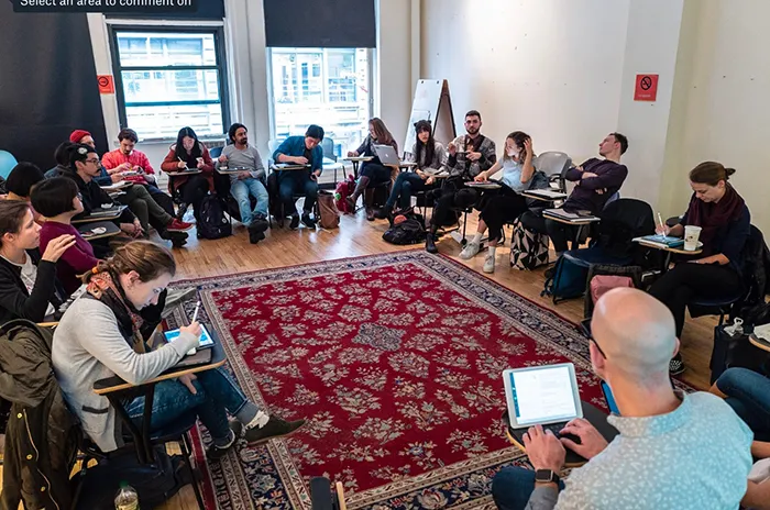 Seventeen people seated along the perimeter of a room—with two large windows, wood floors, and a large area rug—facing a man with his back to the camera.