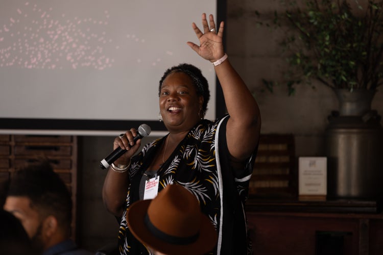 A photograph of a Black woman with short hair, smiling and standing with her back to a presentation screen. She's wearing a black dress with a white print, holding a microphone with her right hand, and raising her left. In the foreground are the backs of the heads of the people in the audience.