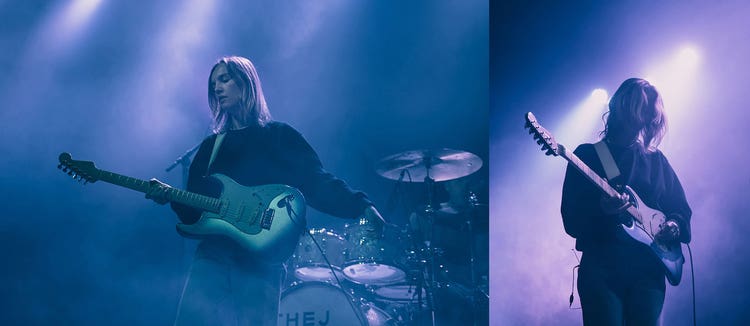 Two photographs of the same white woman with shoulder length blonde hair wearing a sweatshirt and a playing a bass guitar. On the left, she's standing in front of a drum set against a background of blue and purple haze and on the right, she's standing with her head bowed looking at her guitar against a smoke-filled background of stage lights and purple smoke.
