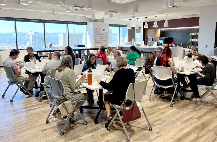 A photograph of a well-lit room, with wall-to-wall windows, wood floors, a small kitchen with a bar-height counter, and a large open space with four square tables (each with four people seated and eating).
