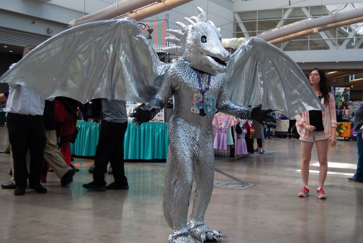 A photograph of a conference hall with attendees and booths in the background. In the foreground is a standing man in a silver-sequined athropomorphized dragon costume with outstretched wings.