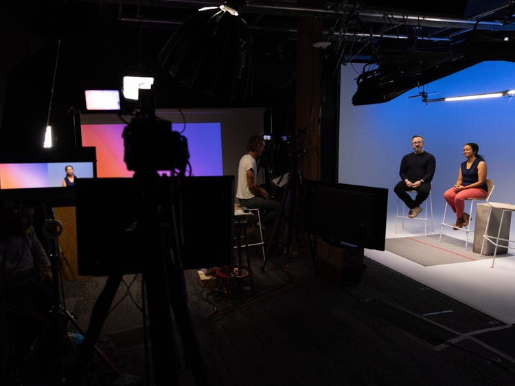 A photograph of a dark room with a cameraman and monitors on the left and a well-lit stage on the right. A white man with glasses, wearing a dark button-down shirt and jeans, and a Southeast Asian woman, wearing red pants and a sleeveless black top, are seated next to each other on the stage.
