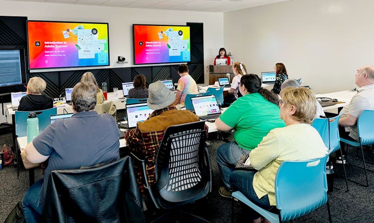 A photograh of twelve people seated at two long conference tables. Their backs are to the camera facing a speaker at the front of the room. Each person has an oen laptop in front of them and there are to large monitors hanging on the wall at the front of the room.
