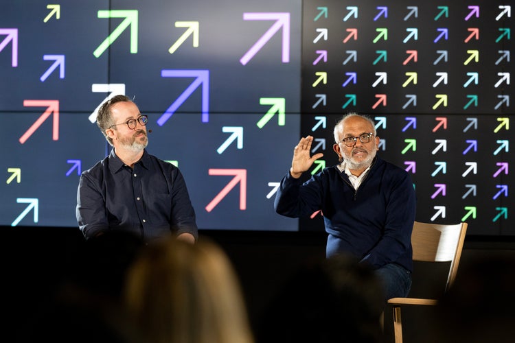Two men, both wearing navy shirts, are seated on stage in front of a grid of colorful arrows pointing diagonally upward to the right. The man on right is raising his right hand as if making a point.