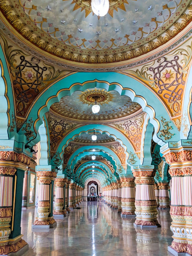 A photograph of a great hall of gilded domes, sculptured pillars and archways, and marble floors that form a grand walkway of pink and teal and gold.