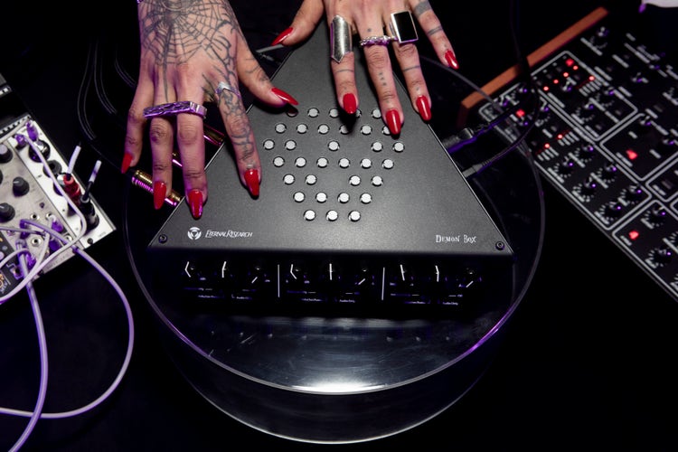 A photograph of tattooed hands with long red nails and chunky rings resting on a black electronic music controller. Knobs, cables, and audio equipment surround the device in low, dramatic lighting that suggests live sound manipulation or experimental music performance.