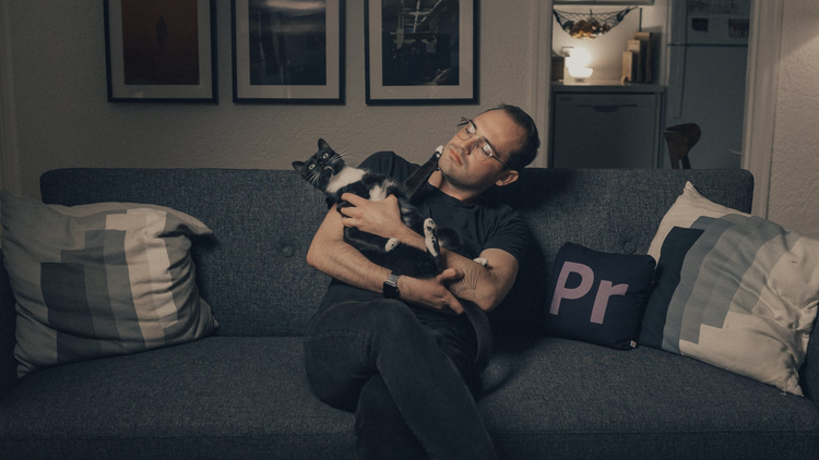 A photograph of a man with glasses sitting on a charcoal grey couch holding a black-and-white cat that's staring at the ceiling.