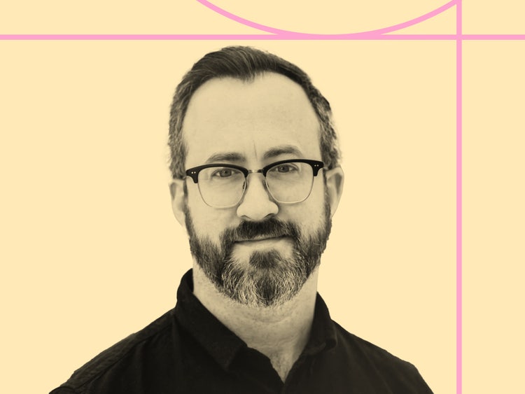 A black-and white photograph of a white man with dark hair, glasses, and a greying mustache and beard on a pale yellow background.