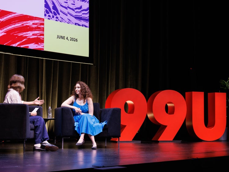 Two women sitting on a stage. The woman on the left is wearing blue pants and has medium wavy brown hair, and the woman on the right has curly brown hair and is wearing a bright blue sun dress. Behind them is a large red 99U cut out.