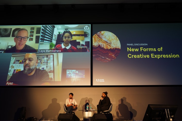 A photograph of two projection screens with two men seated at a small table in front and beneath them: On the left screen are three individuals each on separate conference call screens and on the right are the words Panel Discussion: New Forms of Creative Expression.