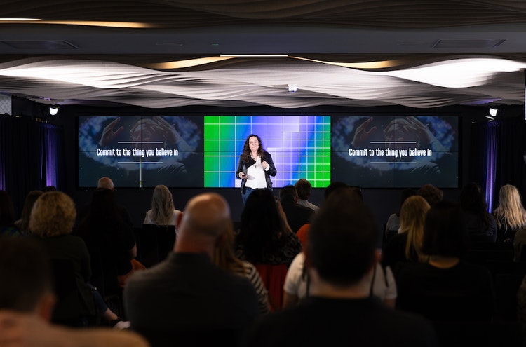 A white woman with dark curly hair, wearing wire-rimmed glasses, a white button-down shirt, and a black jacket, stands speaking in front of screen that reads "Commit to the thing you believe in."