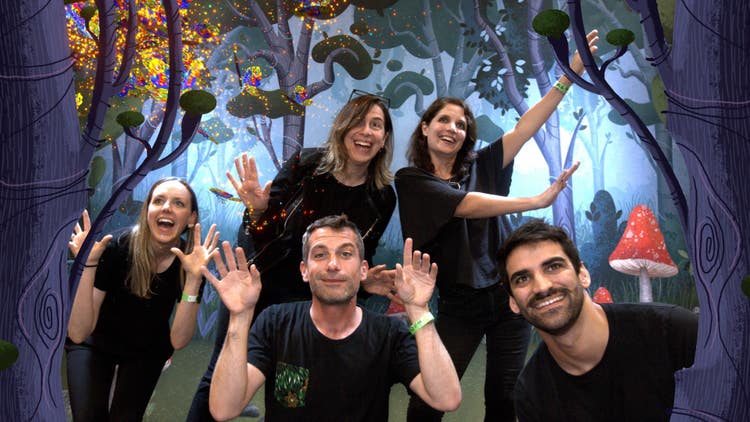 A photograph of two men (in the foreground) and three women (in the background), all wearing black T-shirts, smiling and waving their hands, against a backdrop of what appears to be an enchanted forest.