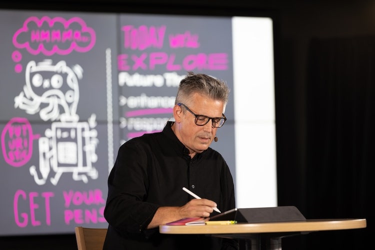 A white man with spiky gray hair, wearing black-rimmed glasses and a black shirt, sits at a table drawing on an iPad with an Apple Pencil, in n front of a screen that reads "Get your pens. Explore."