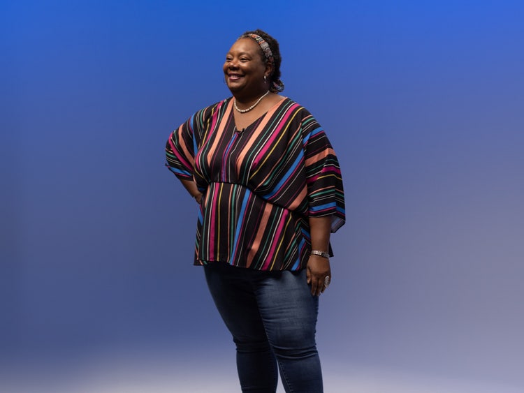 A photograph of a Black woman wearing jeans and a vibrant peach, pink, blue, yellow, and black striped top standing in front of a blue-screen backdrop.