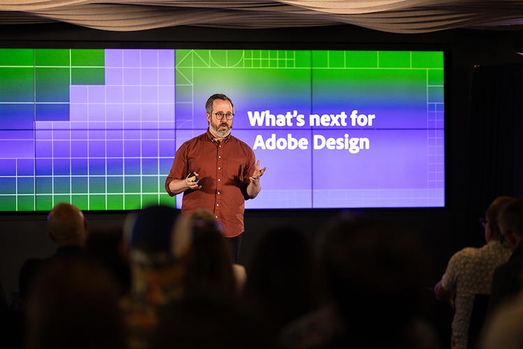 A white man, with dark hair and a beard, wearing black-rimmed glasses and a rust-colored button down shirt stands speaking in front of a screen that reads "What's next for Adobe Design."