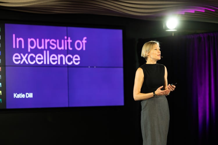 A white woman with short blond hair, wearing a grey skirt and black sleeveless T-shirt, stands on stage holding a device, possibly a remote or microphone, in front of a large screen displaying the text "In pursuit of excellence" just above her name (Katie Dill).
