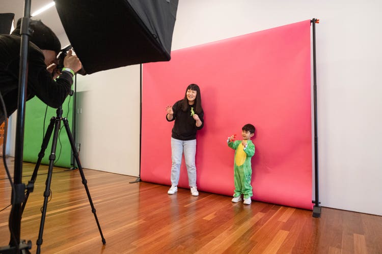 A photograph of a little boy, wearing a green and yellow dinosaur costume, and his mom. Both are standing in the background against a pink screen, and holding plastic dinosaurs, while a photographer in the foreground (using a camera on a tripod) takes their photo.