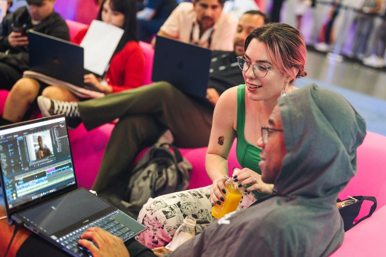 Several people working on laptops in a casual, open space. Two people in the foreground are seated on bright pink lounge furniture, collaborating on video editing. One person looks on while the other demonstrates a feature.