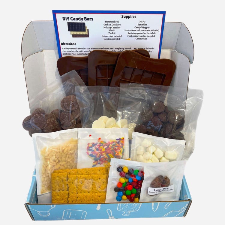 A blue-and-white cardboard shipping box overflowing with supplies for making chocolate bars: milk, white, and dark chocolate chips, almonds, sprinkles, candy-shelled chocolate, marshmallows, and graham crackers.