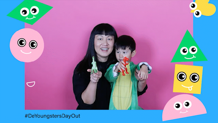 A GIF of a little boy, wearing a green and yellow dinosaur costume, and his mom. Both are against a pink screen background and holding plastic dinosaurs. The GIF is framed in blue and adorned with happy faces on shapes (a heart, diamond, circle, triangle, and square). #DeYoungstersDayOut is in black type in the lower left hand corner.