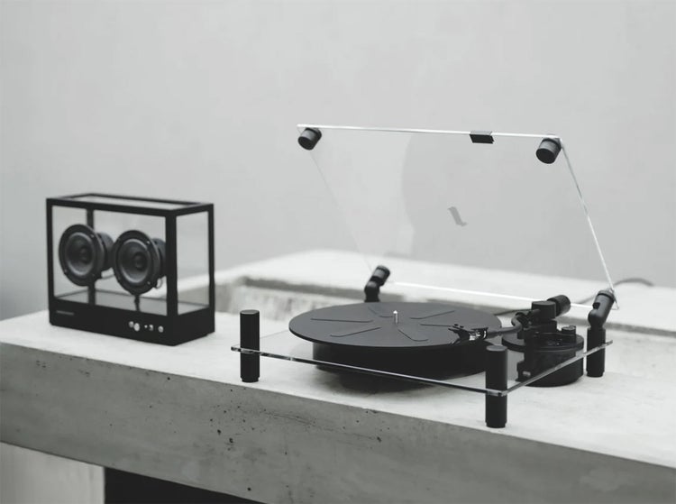 A minimalist photograph of a modern turntable with a clear acrylic dust cover sitting on a concrete surface. A small cube speaker with visible drivers stands beside it. The neutral gray setting emphasizes clean lines, precision, and contemporary audio design.