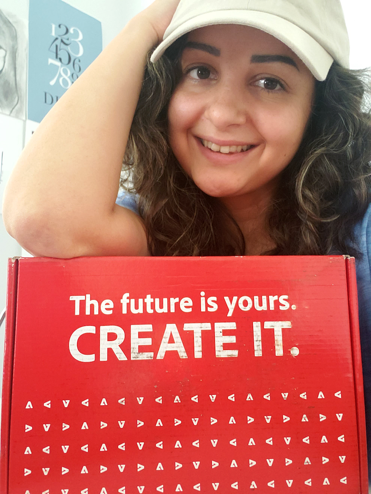 A young woman with curly brown hair and wearing a baseball cap, smiles while leaning forward on a red cardboard box that reads "The future is yours, CREATE IT,"