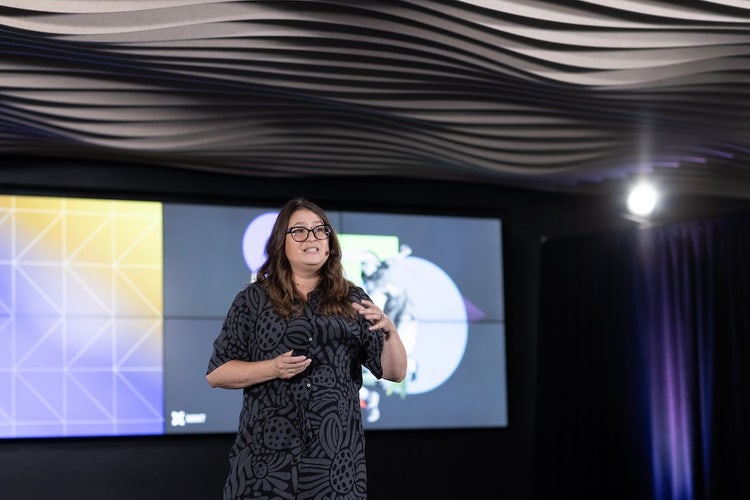 A white woman, with dark hair, wearing black-rimmed glasses and a gray-and-black patterened jumpsuit stands speaking in front of a screen.