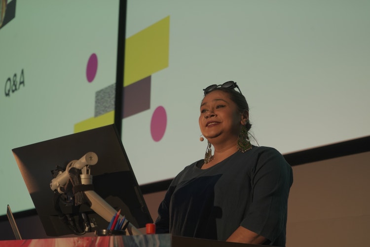 A photograph of a woman, wearing large, ornate earrings and sunglasses perched on her head. She's standing behind a podium with a computer screen on it and behind her are two large projection screens