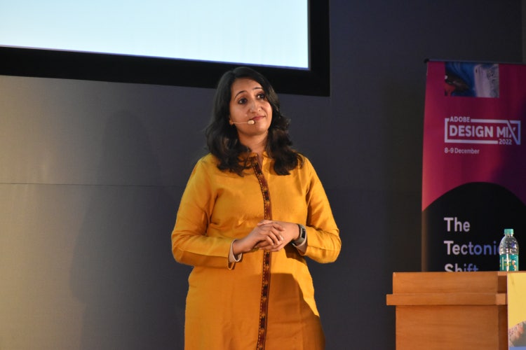 A photograph of a woman with long dark hair and, wearing an orange coat standing in front of a large projection screen. To her right is a podium and.banner with the Design Mix logotype in white on a pink background.