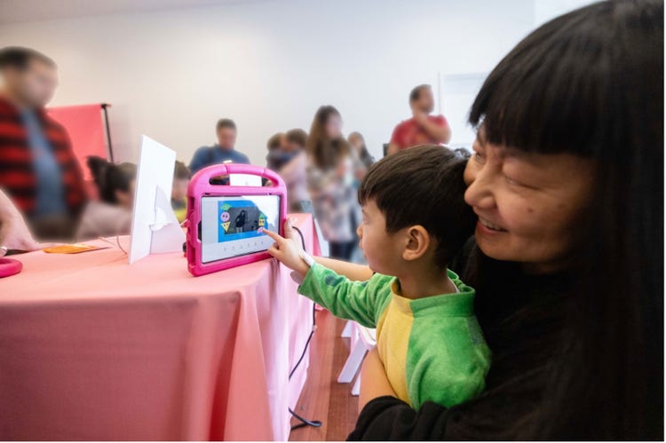 A photograph of a little boy, wearing a green and yellow dinosaur costume, sitting on his mom's lap at a table with a pink tablecloth. People are standing in the background as his mom watches over his shoulder while he uses an application on an iPad.