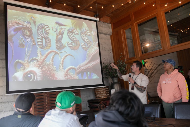A photograph of a white man (wearing a white T-shirt and holding a microphone) and a Black woman (wearing jeans, a peach hoodie, and blue baseball cap). Both of them are facing an audience (whose heads are visible in the foreground) and the man is speaking and pointing to a large presentation screen with a friendly-looking sea creature and the word ABYSS on it.