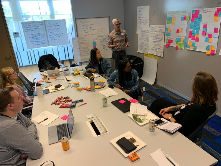 A conference room with a man stading at a white board ready to take notes while another man and two women seated around a conference table (with iPads, laptop computers, water glasses and bttles, and sticky notes) appear to be looking at someone outside the camera view.