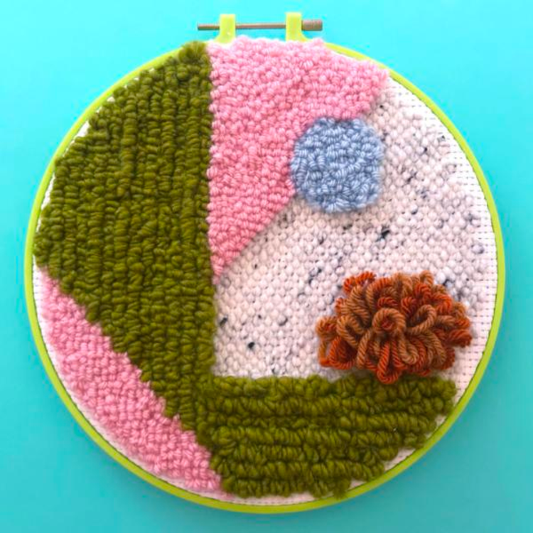 An embroidery hoop with a completed punch needle project comprise of yarn colors of olive green, rust orange, pink, and light blue in a geometric pattern.