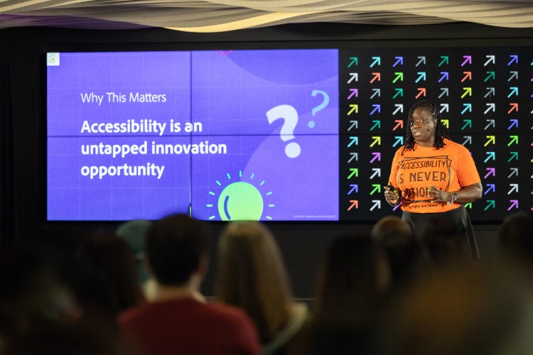 A Black woman with her dark hair in twists and wearing an orange that reads "Accessibility Is Never Optional" stands in front of an audience giving a presentation. Behind her is a screen divided in two sections. The one on the left reads, "Why This Matters: Accessibility is an untapped innovation opportunity'" and the one on the right a grid of colorful arrows pointing diagonally upward to the right.