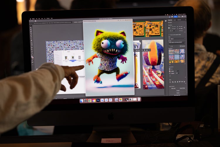 A photograph of a computer screen with a finger pointing at it. On the screen is a digital image of a friendly-looking lime green-furred monster with bulging eyes, sharp teeth, and cat-like ears, wearing a printed T-shirt. He appears to be trying to walk off the screen.