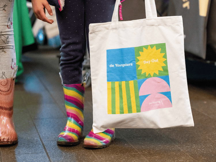 A photograph of the legs of two children both wearing multicolored rainboots. The child on the right is holding a canvas bag with with a blue, green, yellow, and pink design that reads de Youngsters Day Out.