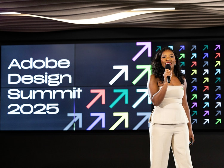 A Black woman with long dark hair, wearing an off-white pantsuit, stands on stage holding a microphone.. Behind her, a large screen displays the words Adobe Design Summit 2025 alongside a grid of colorful arrows pointing diagonally upward to the right.